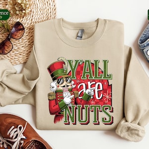 May include: Beige crewneck sweatshirt with a graphic of a nutcracker wearing sunglasses and a military-style uniform. The text "Y'ALL are NUTS" is displayed in green and red. The sweatshirt is paired with blue jeans and brown shoes.