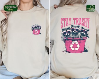 May include: Beige sweatshirt with a graphic of three raccoons wearing sunglasses in a pink trash can. The text "STAY TRASHY" is above the raccoons on the back. A smaller version of the graphic is on the front left chest.
