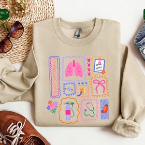 May include: Beige sweatshirt with a colorful graphic design featuring medical-themed illustrations. The design includes pink lungs, a stethoscope, and the text "RESPIRATORY THERAPIST" in a rectangular frame. The sweatshirt is paired with brown shoes and sunglasses.
