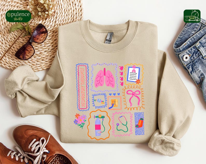 May include: Beige sweatshirt with a colorful graphic design featuring medical-themed illustrations. The design includes pink lungs, a stethoscope, and the text "RESPIRATORY THERAPIST" in a rectangular frame. The sweatshirt is paired with brown shoes and sunglasses.