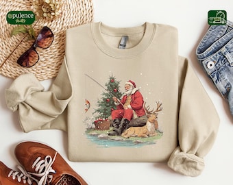 May include: Beige sweatshirt featuring a festive illustration of Santa Claus fishing near a decorated Christmas tree. Santa is wearing a red suit and hat, with a deer resting nearby. The design includes a fishing rod and a caught fish.