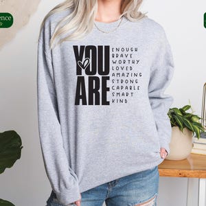 Puede incluir: Sudadera gris con las palabras "YOU ARE" en letras negras grandes. Las palabras "ENOUGH, BRAVE, WORTHY, LOVED, AMAZING, STRONG, CAPABLE, SMART, KIND" est&aacute;n dispuestas en una columna vertical a la derecha de las palabras "YOU ARE".