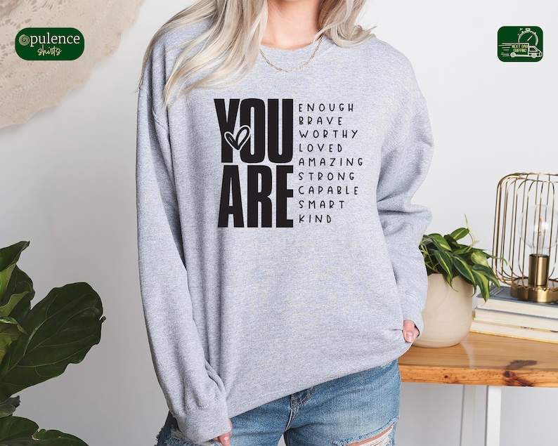 Puede incluir: Sudadera gris con las palabras "YOU ARE" en letras negras grandes. Las palabras "ENOUGH, BRAVE, WORTHY, LOVED, AMAZING, STRONG, CAPABLE, SMART, KIND" est&aacute;n dispuestas en una columna vertical a la derecha de las palabras "YOU ARE".