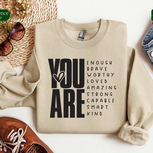 Puede incluir: Una sudadera beige con las palabras "YOU ARE" en letras negras grandes. Las palabras "ENOUGH, BRAVE, WORTHY, LOVED, AMAZING, STRONG, CAPABLE, SMART, KIND" est&aacute;n dispuestas alrededor de las palabras "YOU ARE" en una columna vertical.