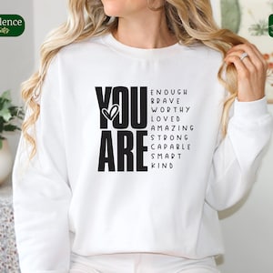 Puede incluir: Sudadera blanca con texto negro que dice "YOU ARE" con un s&iacute;mbolo de coraz&oacute;n y una lista de palabras positivas que incluyen "enough, brave, worthy, loved, amazing, strong, capable, smart, kind".