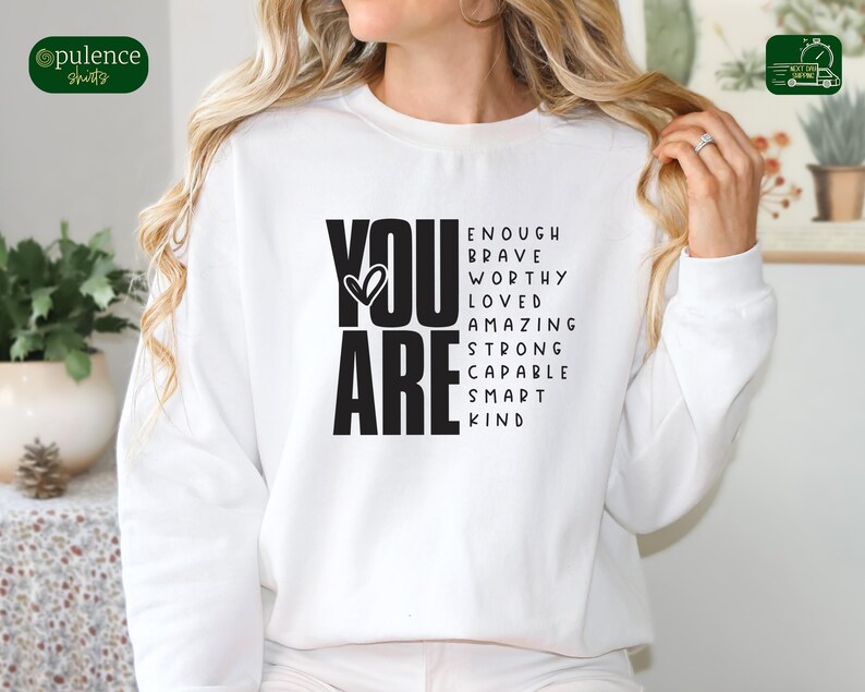 Puede incluir: Sudadera blanca con texto negro que dice "YOU ARE" con un s&iacute;mbolo de coraz&oacute;n y una lista de palabras positivas que incluyen "enough, brave, worthy, loved, amazing, strong, capable, smart, kind".