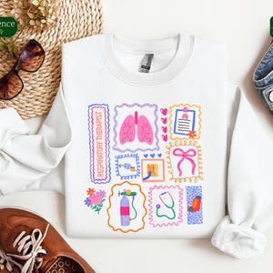 May include: White sweatshirt with colorful illustrations related to respiratory therapy. The design includes images of lungs, a stethoscope, and the text "RESPIRATORY THERAPIST" in a rectangular frame. The sweatshirt is paired with sunglasses and brown shoes.