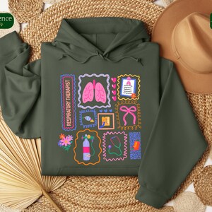 May include: Dark green hoodie with a colorful graphic design featuring medical-themed illustrations. The design includes images of lungs, a stethoscope, and a bow, with the text "RESPIRATORY THERAPIST".