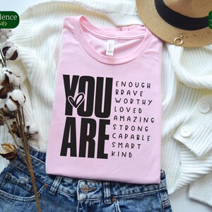 Puede incluir: Camiseta rosa con texto negro que dice "YOU ARE" y una lista de palabras positivas que incluyen "enough", "brave", "worthy", "loved", "amazing", "strong", "capable", "smart" y "kind".