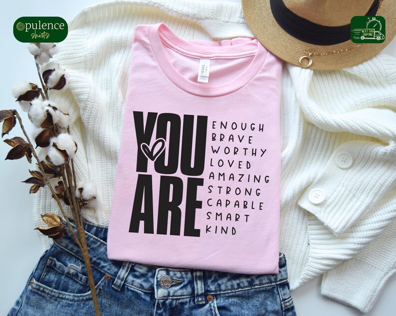 Puede incluir: Camiseta rosa con texto negro que dice "YOU ARE" y una lista de palabras positivas que incluyen "enough", "brave", "worthy", "loved", "amazing", "strong", "capable", "smart" y "kind".