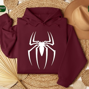 Puede incluir: Una sudadera con capucha color burdeos con un logotipo de Spiderman blanco en la parte delantera. La sudadera est&aacute; colocada sobre una superficie tejida con un sombrero marr&oacute;n y un abanico.
