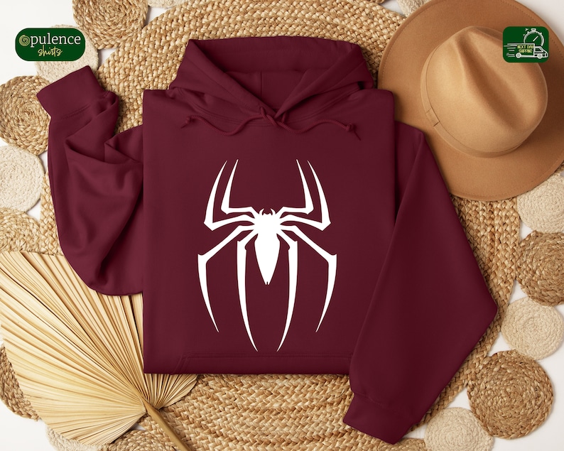 Puede incluir: Una sudadera con capucha color burdeos con un logotipo de Spiderman blanco en la parte delantera. La sudadera est&aacute; colocada sobre una superficie tejida con un sombrero marr&oacute;n y un abanico.