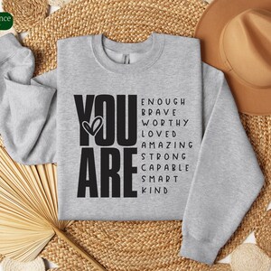 Puede incluir: Sudadera gris con las palabras "YOU ARE" en letras negras grandes. Las palabras "ENOUGH, BRAVE, WORTHY, LOVED, AMAZING, STRONG, CAPABLE, SMART, KIND" est&aacute;n dispuestas alrededor de las palabras "YOU ARE" en una nube de palabras.