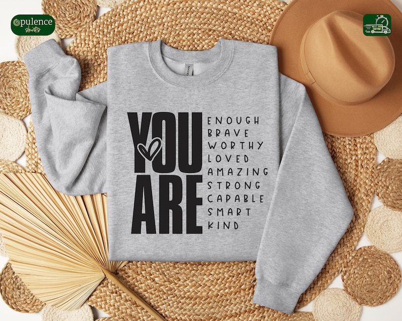 Puede incluir: Sudadera gris con las palabras "YOU ARE" en letras negras grandes. Las palabras "ENOUGH, BRAVE, WORTHY, LOVED, AMAZING, STRONG, CAPABLE, SMART, KIND" est&aacute;n dispuestas alrededor de las palabras "YOU ARE" en una nube de palabras.