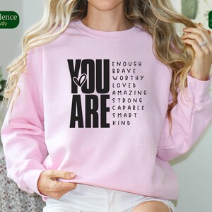 Puede incluir: Sudadera rosa con las palabras "YOU ARE" en negro, rodeadas de una lista de afirmaciones positivas que incluyen "enough", "brave", "worthy", "loved", "amazing", "strong", "capable", "smart" y "kind".