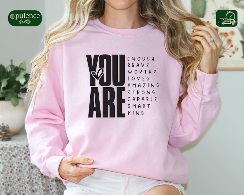Puede incluir: Sudadera rosa con las palabras "YOU ARE" en negro, rodeadas de una lista de afirmaciones positivas que incluyen "enough", "brave", "worthy", "loved", "amazing", "strong", "capable", "smart" y "kind".