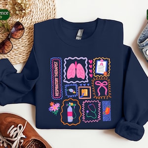 May include: Navy blue sweatshirt featuring a colorful graphic design. The design includes illustrations of lungs, a stethoscope, and other medical-related images, along with the text "Respiratory Therapist". The sweatshirt is a crew neck style.