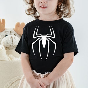 Puede incluir: Un ni&ntilde;o o ni&ntilde;a lleva una camiseta negra con un gr&aacute;fico de ara&ntilde;a blanca. La ara&ntilde;a es un dise&ntilde;o estilizado de una telara&ntilde;a con una ara&ntilde;a en el centro.