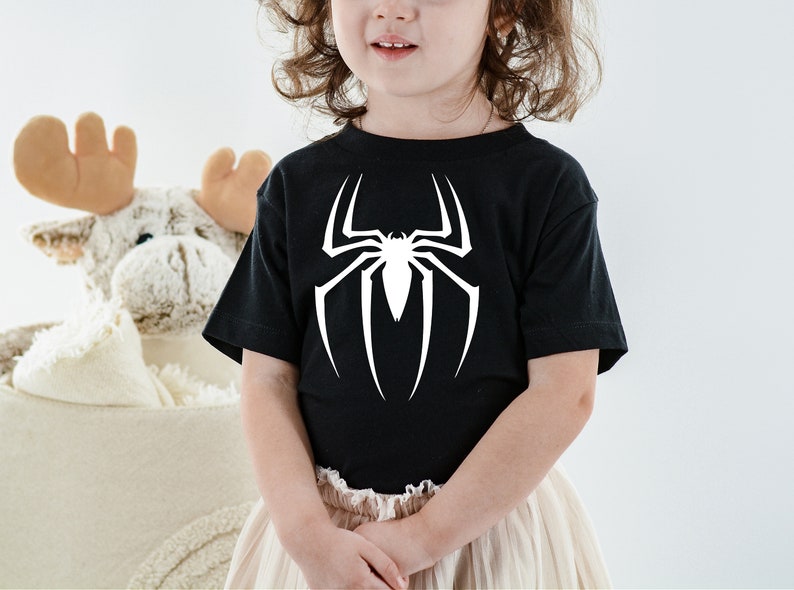 Puede incluir: Un ni&ntilde;o o ni&ntilde;a lleva una camiseta negra con un gr&aacute;fico de ara&ntilde;a blanca. La ara&ntilde;a es un dise&ntilde;o estilizado de una telara&ntilde;a con una ara&ntilde;a en el centro.