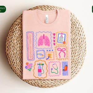 May include: A light pink t-shirt with a colorful graphic design featuring medical-themed illustrations. The design includes images of lungs, a stethoscope, and a respiratory therapist label. The shirt is folded on a woven wicker mat.