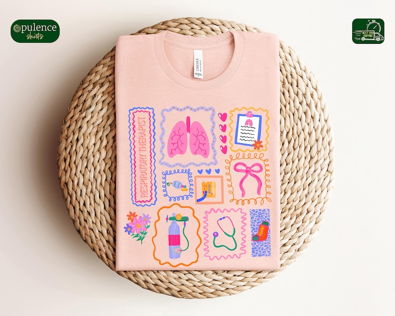 May include: A light pink t-shirt with a colorful graphic design featuring medical-themed illustrations. The design includes images of lungs, a stethoscope, and a respiratory therapist label. The shirt is folded on a woven wicker mat.