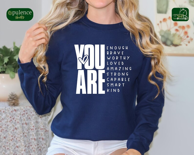 Puede incluir: Sudadera azul marino con texto blanco que dice "YOU ARE" con las palabras "enough, brave, worthy, loved, amazing, strong, capable, smart, kind" dispuestas alrededor de las palabras "YOU ARE".