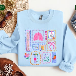 May include: A light blue sweatshirt featuring various medical-themed illustrations, including lungs, a stethoscope, and a medical chart. The words "RESPIRATORY THERAPIST" are printed on the left side. The design is framed by decorative borders. The sweatshirt is made of a soft material.