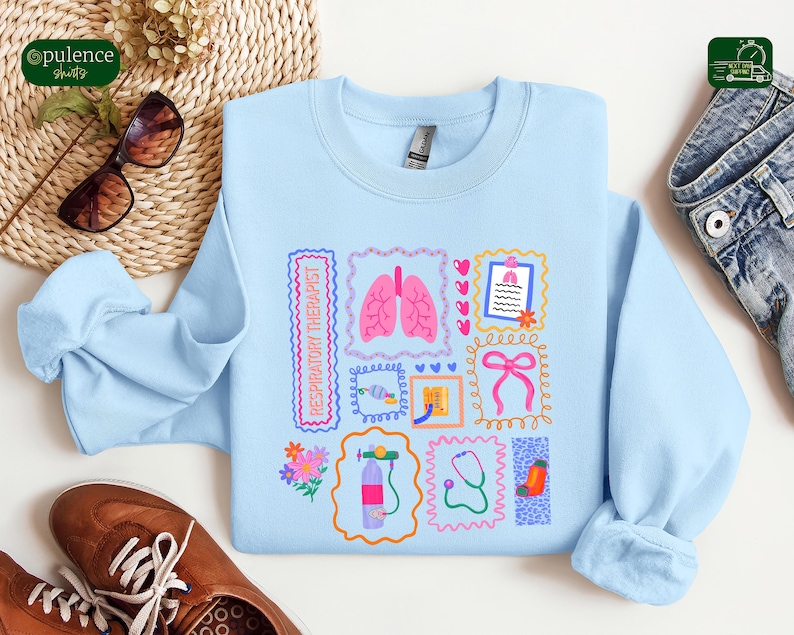 May include: A light blue sweatshirt featuring various medical-themed illustrations, including lungs, a stethoscope, and a medical chart. The words "RESPIRATORY THERAPIST" are printed on the left side. The design is framed by decorative borders. The sweatshirt is made of a soft material.