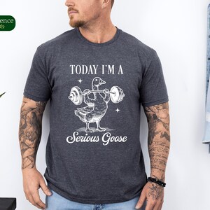 May include: A dark heather grey t-shirt with a white graphic of a goose lifting weights. The text "Today I'm a Serious Goose" is printed below the graphic.