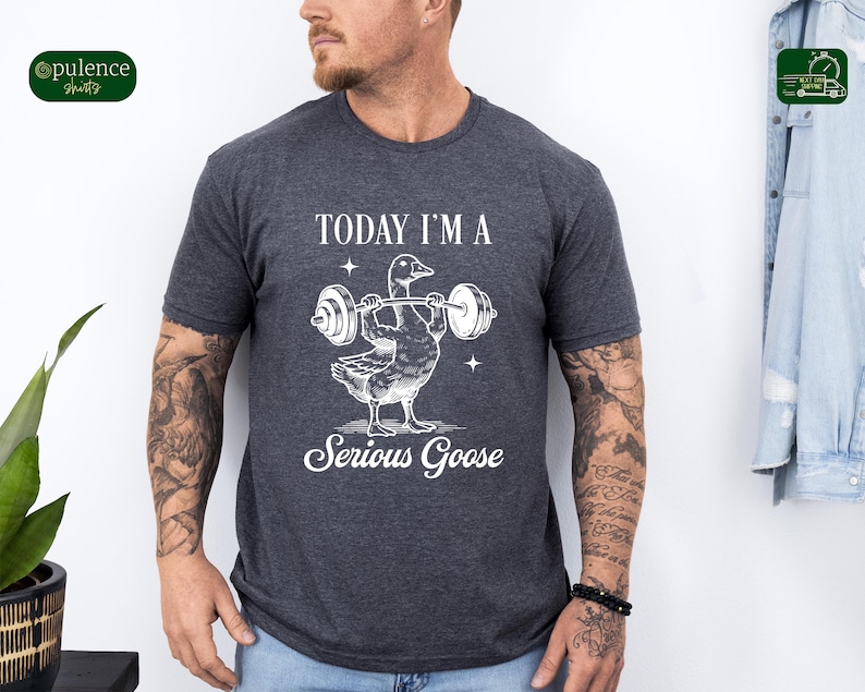 May include: A dark heather grey t-shirt with a white graphic of a goose lifting weights. The text "Today I'm a Serious Goose" is printed below the graphic.