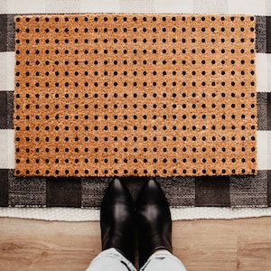 May include: A rectangular coir doormat featuring a repeating pattern of small, solid black polka dots across its natural tan, fibrous surface. The mat is positioned horizontally, showcasing a consistent grid of dark circular markings.