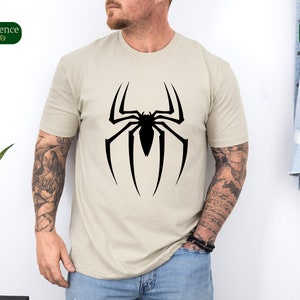 Puede incluir: Una camiseta beige con un gr&aacute;fico de ara&ntilde;a negra. La ara&ntilde;a est&aacute; en un dise&ntilde;o de telara&ntilde;a.