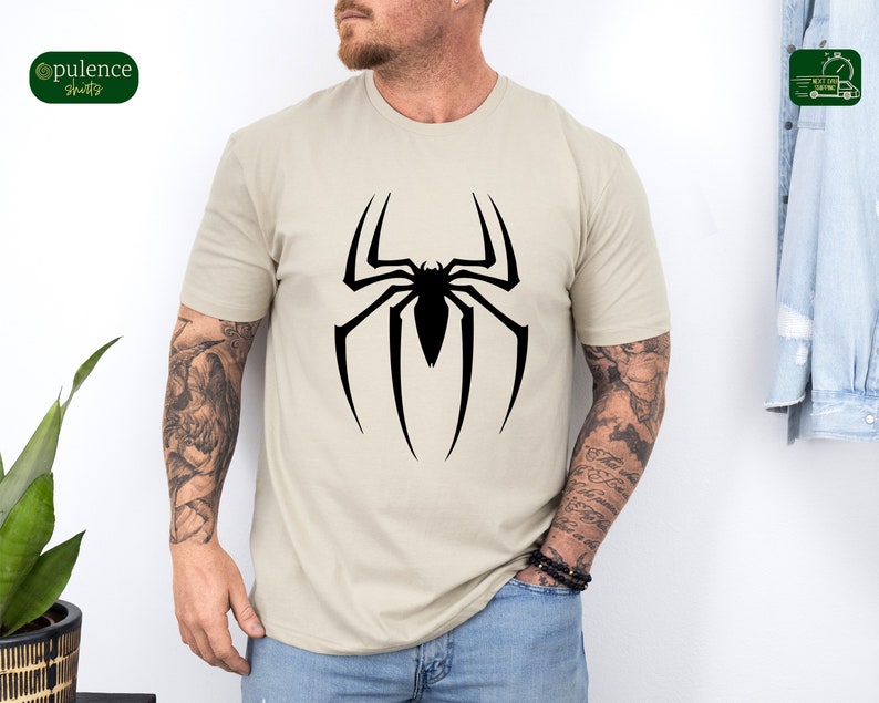 Puede incluir: Una camiseta beige con un gr&aacute;fico de ara&ntilde;a negra. La ara&ntilde;a est&aacute; en un dise&ntilde;o de telara&ntilde;a.