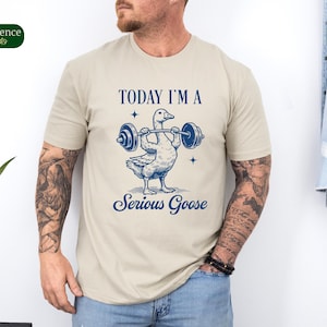 May include: A beige t-shirt with a graphic of a goose lifting a barbell. The text "TODAY I'M A SERIOUS GOOSE" is printed on the shirt.
