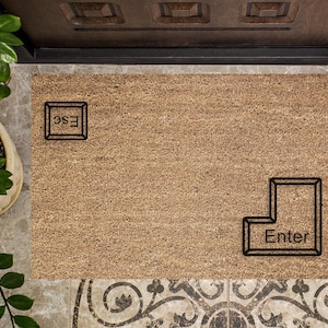 May include: A rectangular doormat made of natural fibers, featuring the words "ESC" and "Enter" in a black, keyboard-inspired design. The mat is placed in front of a dark wooden door.