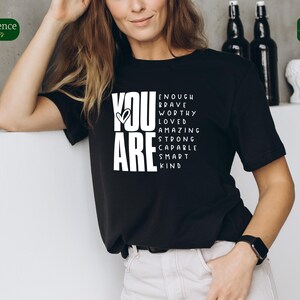 Puede incluir: Camiseta negra con texto blanco que dice "You Are" con las palabras "Enough, Brave, Worthy, Loved, Amazing, Strong, Capable, Smart, Kind" dispuestas alrededor de las palabras "You Are".