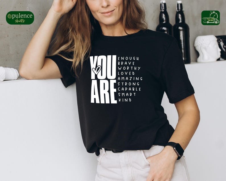 Puede incluir: Camiseta negra con texto blanco que dice "You Are" con las palabras "Enough, Brave, Worthy, Loved, Amazing, Strong, Capable, Smart, Kind" dispuestas alrededor de las palabras "You Are".