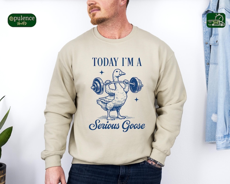 May include: A beige sweatshirt with a blue graphic of a goose lifting a barbell. The text "TODAY I'M A SERIOUS GOOSE" is printed on the sweatshirt.