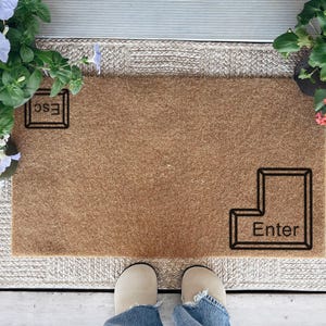 May include: A rectangular brown coir doormat with the text "Esc" and "Enter" in black, keyboard key outlines. The mat is surrounded by a woven border and flanked by potted flowers in shades of blue, white, yellow, and pink.