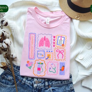 May include: A light pink t-shirt featuring a collage of medical-themed illustrations, including lungs, a stethoscope, and a bow. The words "RESPIRATORY THERAPIST" are printed vertically. The shirt is folded on top of blue jeans and a white sweater.