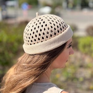 May include: A beige knitted hat with a perforated design and a small, rounded top. The hat has a ribbed brim and a slightly domed crown. The hat is made of a light-colored yarn and appears to be handmade.