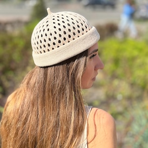 May include: A beige knit hat with a perforated design and a small, rounded top. The hat has a folded brim and is worn by a person with long, light brown hair. The background is blurred, suggesting an outdoor setting.