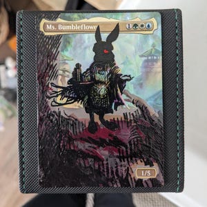 May include: A trading card featuring a black rabbit-like figure with a red eye, wearing a patterned outfit. The card is labeled "Ms. Bumbleflowe" and includes mana symbols. The card is in a black card holder with green stitching.