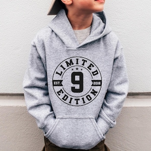 May include: A gray hooded sweatshirt with a black circle graphic that says "Limited Edition" and has a number "9" inside the circle. The sweatshirt is being worn by a child wearing a beige baseball cap and blue sunglasses.