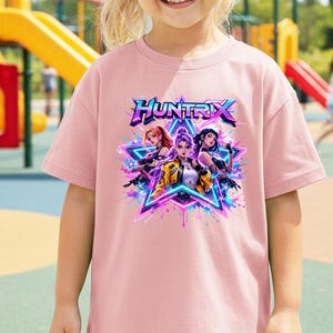 May include: A pale pink t-shirt with the word "HUNTRIX" in a colourful font. The graphic shows three female characters in a star design with neon blue and purple accents. The t-shirt is being worn by a young child.