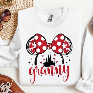 K&ouml;nnte beinhalten: Wei&szlig;er Pullover mit einem roten und wei&szlig;en Minnie Mouse Ohren-Design, einer schwarzen Silhouette von Schloss Aschenputtel und dem Wort "granny" in roten Buchstaben.