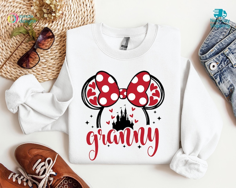 K&ouml;nnte beinhalten: Wei&szlig;er Pullover mit einem roten und wei&szlig;en Minnie Mouse Ohren-Design, einer schwarzen Silhouette von Schloss Aschenputtel und dem Wort "granny" in roten Buchstaben.