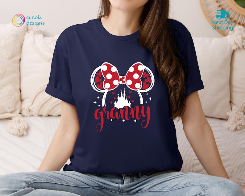 K&ouml;nnte beinhalten: Ein marineblaues T-Shirt mit einer wei&szlig;en Grafik eines roten und wei&szlig;en gepunkteten Bogens, eines wei&szlig;en Schlosses und dem Wort "granny" in Wei&szlig; mit roten Akzenten.