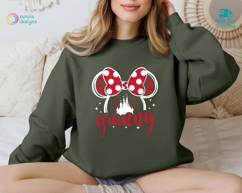 K&ouml;nnte beinhalten: Gr&uuml;ner Pullover mit einem wei&szlig;en Umriss von Mickey Mouse Ohren mit einer roten Schleife und dem Wort "granny" in roten Buchstaben.