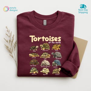May include: Burgundy sweatshirt featuring the text "Tortoises of the World" above illustrations of various tortoise species. The sweatshirt has a crew neck and long sleeves. The design includes the names of each tortoise species.